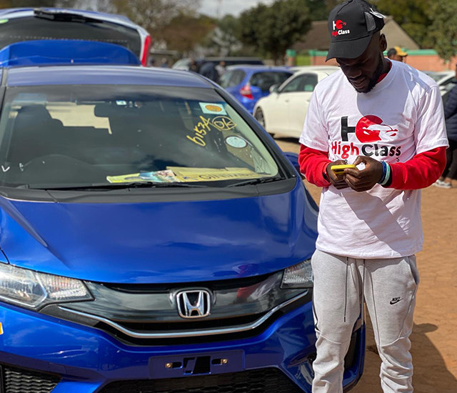 A salesman checking his phone beside a blue Honda Fit on the forecourt